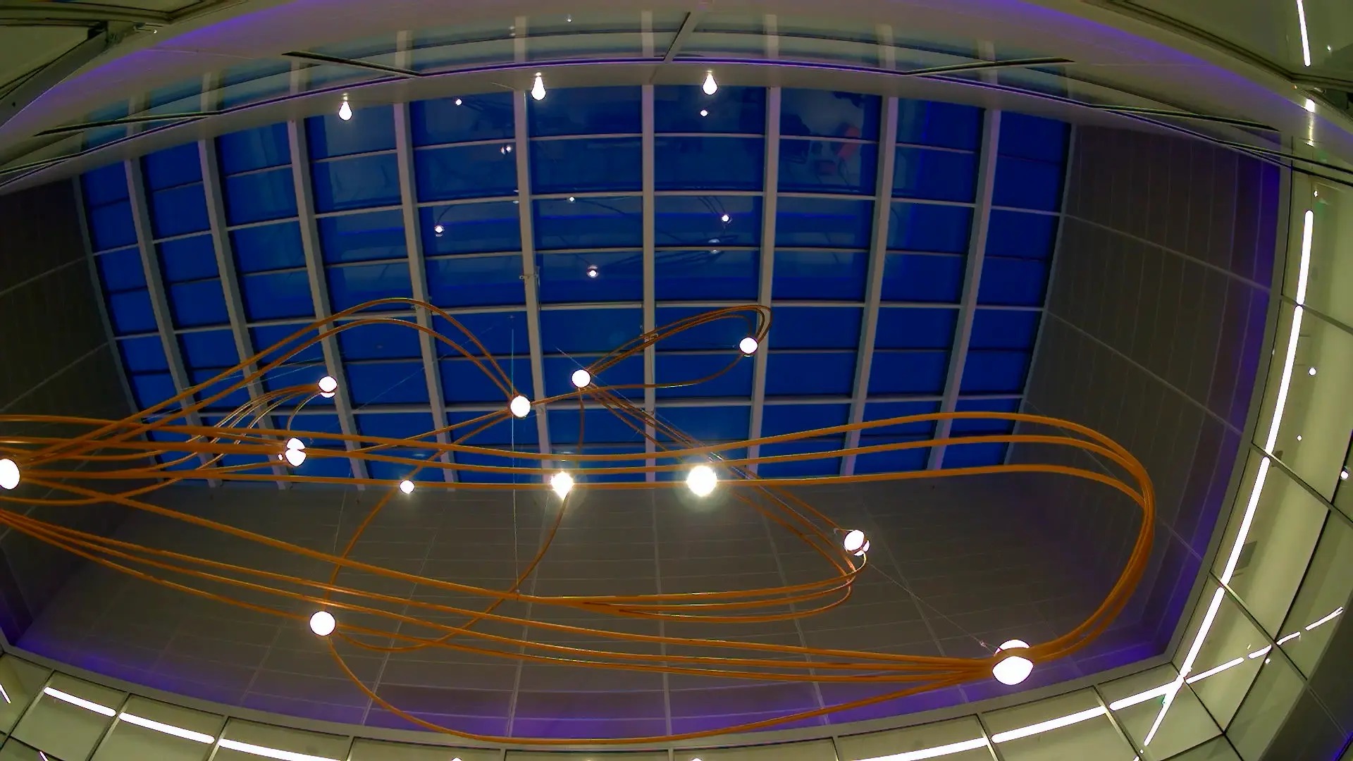Suspended copper-tube light sculpture beneath a blue glass atrium ceiling
