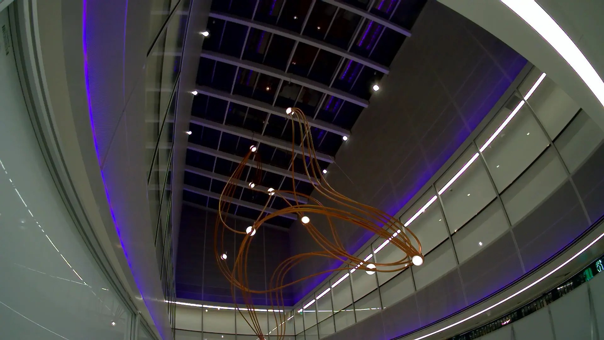 Suspended copper-tube light sculpture in a curved airport atrium washed with violet light